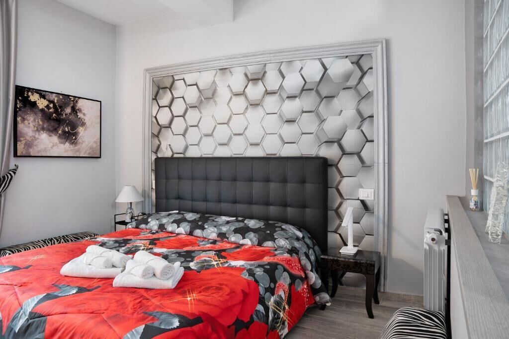 A modern hotel room with a geometric-patterned headboard, a neatly made bed with a red and black floral bedspread, and a side table with a lamp.