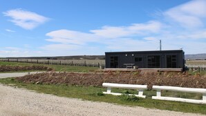 Un bâtiment moderne doté d’un toit plat et de grandes fenêtres, entouré d’une zone délimitée par une clôture, avec un chemin de gravier devant.
