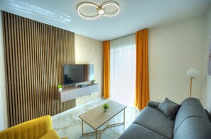 Een moderne woonkamer met een flatscreen televisie bevestigd aan een houten paneelwand, een grijze bank, een gele fauteuil en een marmeren vloer.