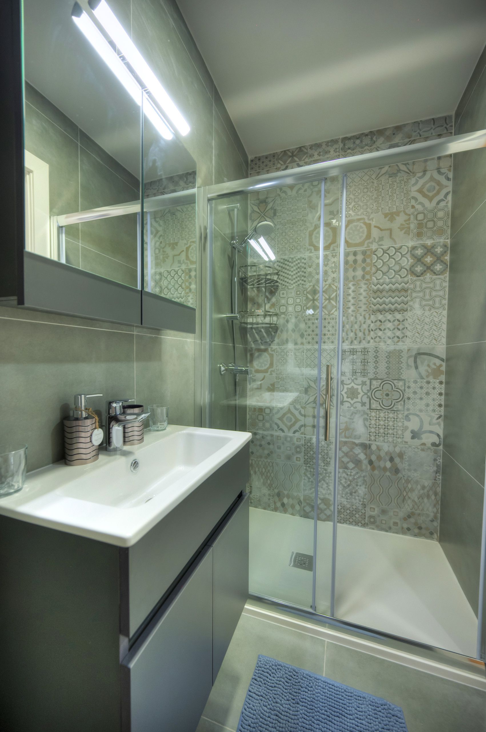 A modern bathroom with a glass shower enclosure, a white sink with a mirror above it, and patterned tiles on the walls.