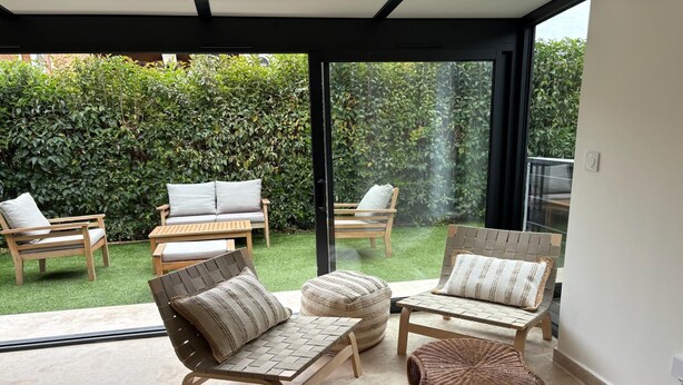 Une terrasse moderne, dotée de meubles en osier, d’une porte vitrée donnant sur un jardin et d’un sol carrelé.