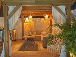 A cozy outdoor seating area with a wooden table, cushioned chairs, and hanging lamps.