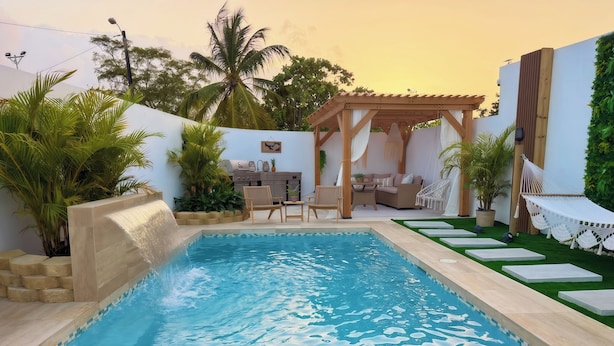 A swimming pool with a waterfall feature, surrounded by outdoor furniture and greenery.