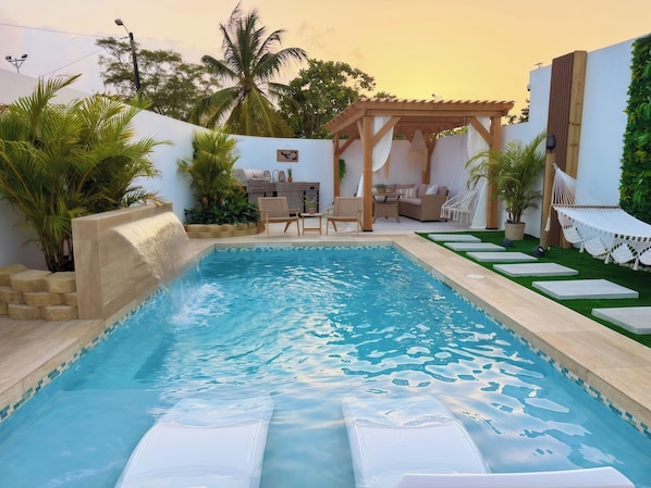 A swimming pool with a waterfall feature, surrounded by outdoor furniture and greenery.