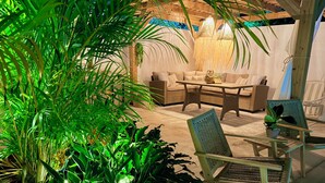 A cozy outdoor seating area with wicker furniture, a hanging light fixture, and lush greenery.