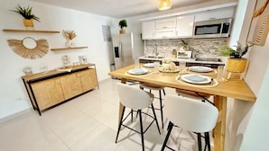 A modern kitchen with white cabinets, a wooden dining table, and bar stools. There are plants on the shelves and a microwave above the counter.