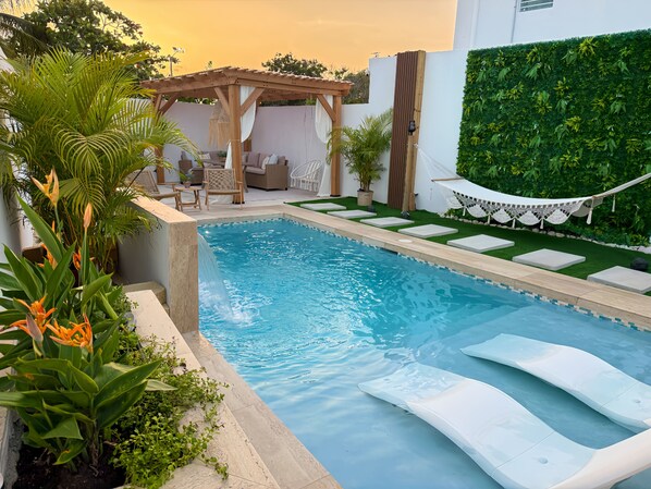 A swimming pool with a waterfall feature, surrounded by a garden with various plants and a hammock.