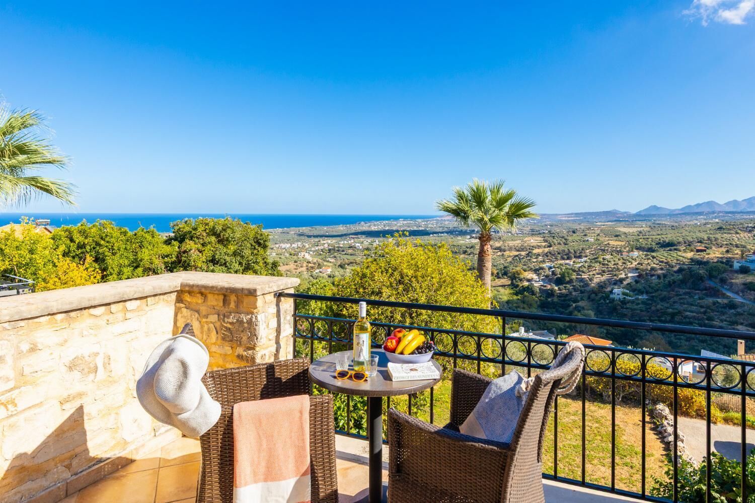 Un balcón con mobiliario de mimbre, una mesa con una botella y fruta, y vista a un paisaje costero.