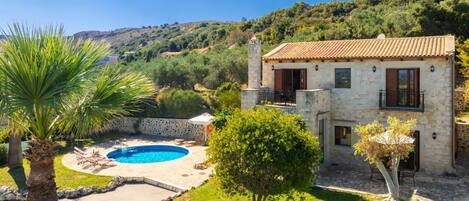 A two-story stone house with a swimming pool, surrounded by lush greenery and hills.