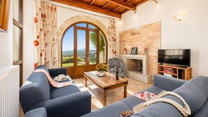 A living room with a fireplace, a television, and a view of the countryside through large windows. - Villa Top Hill - Three Bedroom Villa, Sleeps 6 (Agia Triada)