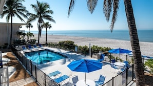 A pool area with blue chairs and umbrellas, a view of the beach, and palm trees. - Beachfront Bliss on Longboat Key- Steps to the sand (Longboat Key)