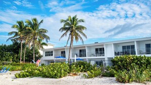 A beachfront hotel with palm trees, a sandy beach, and a white building with multiple windows and balconies. - Beachfront Bliss on Longboat Key- Steps to the sand (Longboat Key)