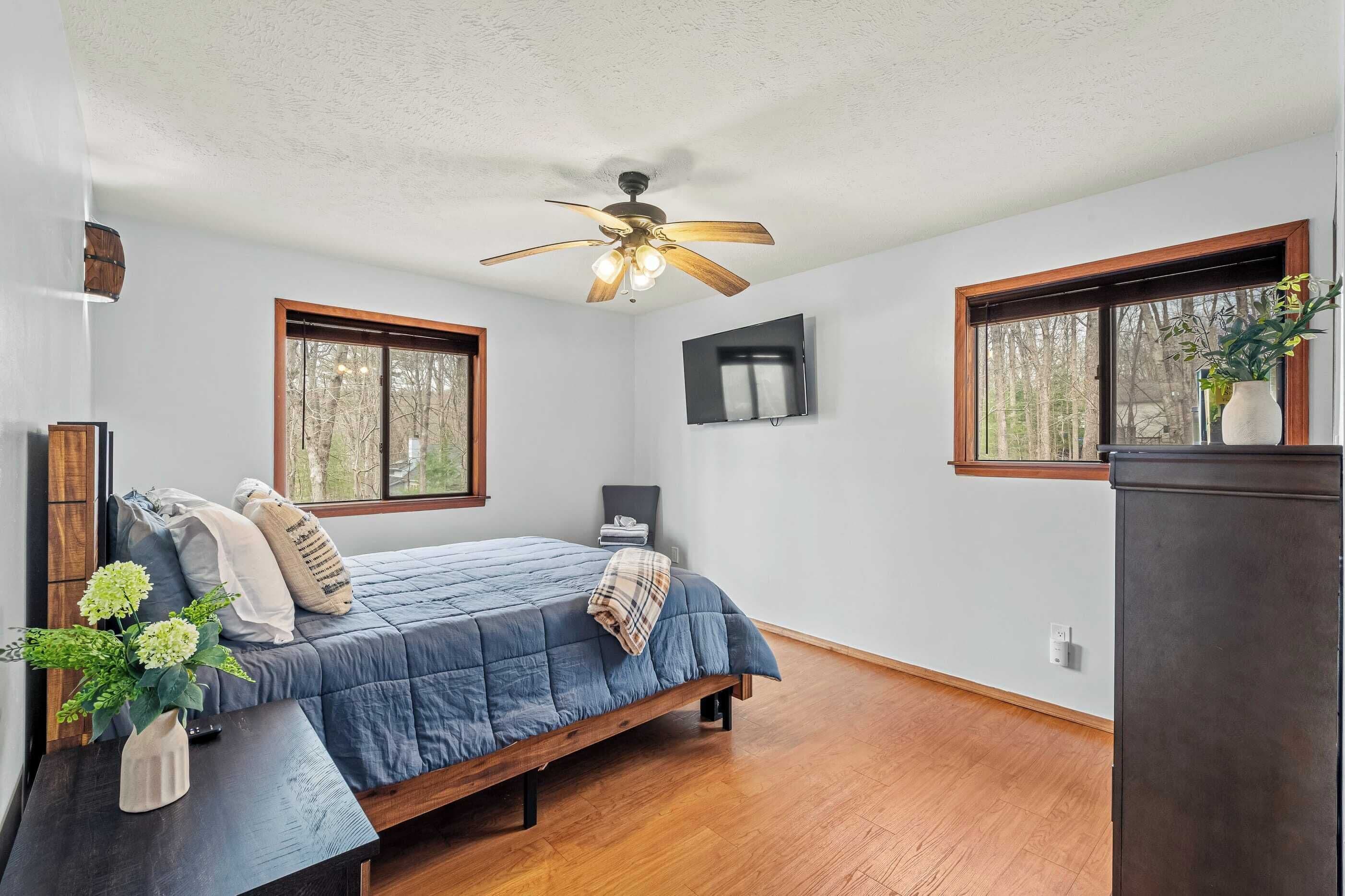 Un dormitorio con cama, ventilador de techo, televisor y ventanas.