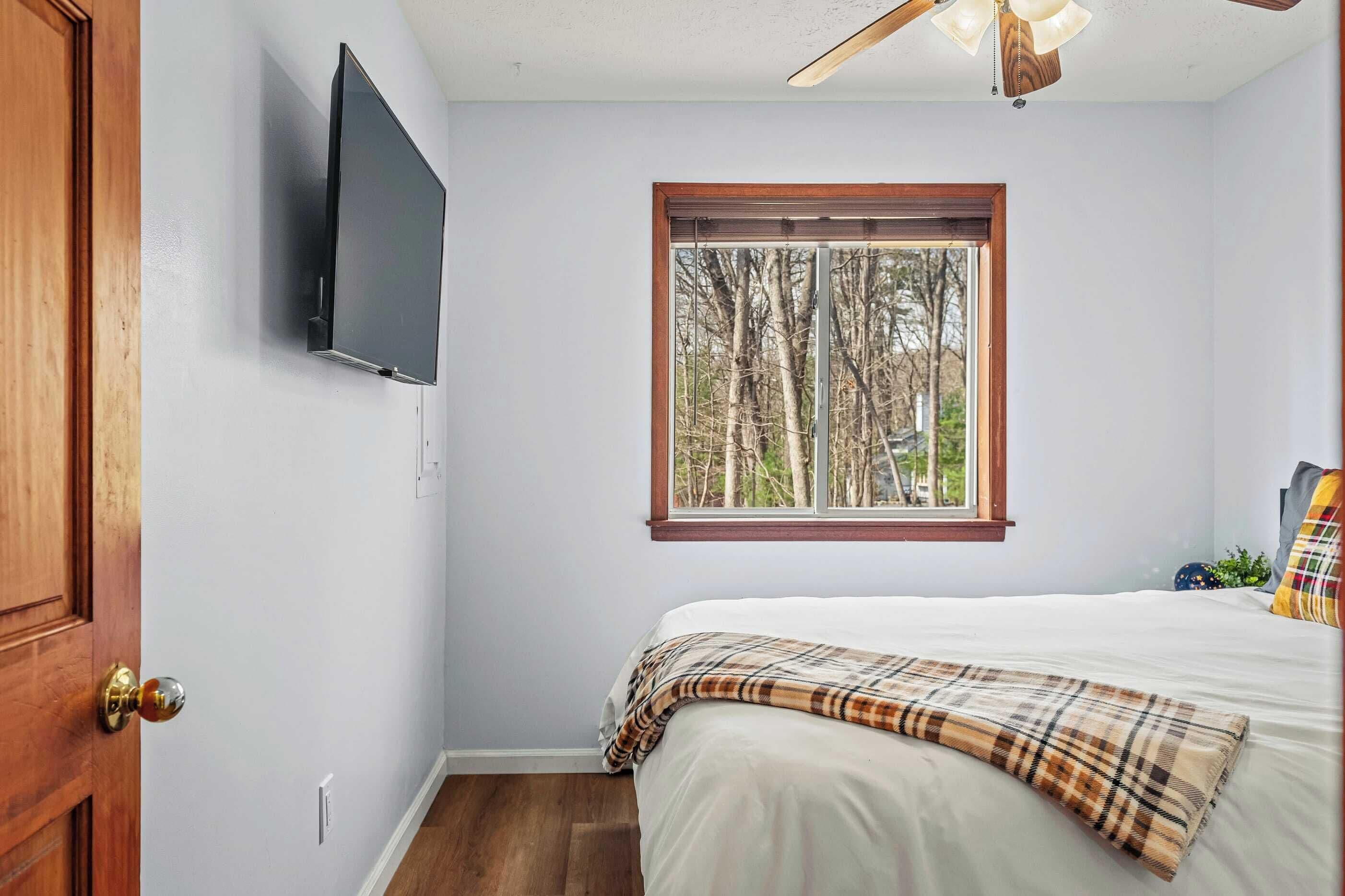 A bedroom with a bed, a television mounted on the wall, a window with a view of trees, and a ceiling fan.