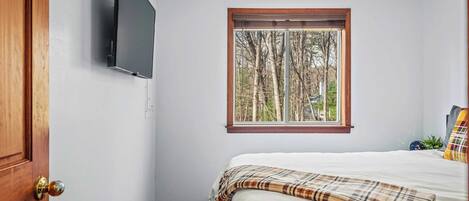 A bedroom with a bed, a television mounted on the wall, a window with a view of trees, and a ceiling fan.