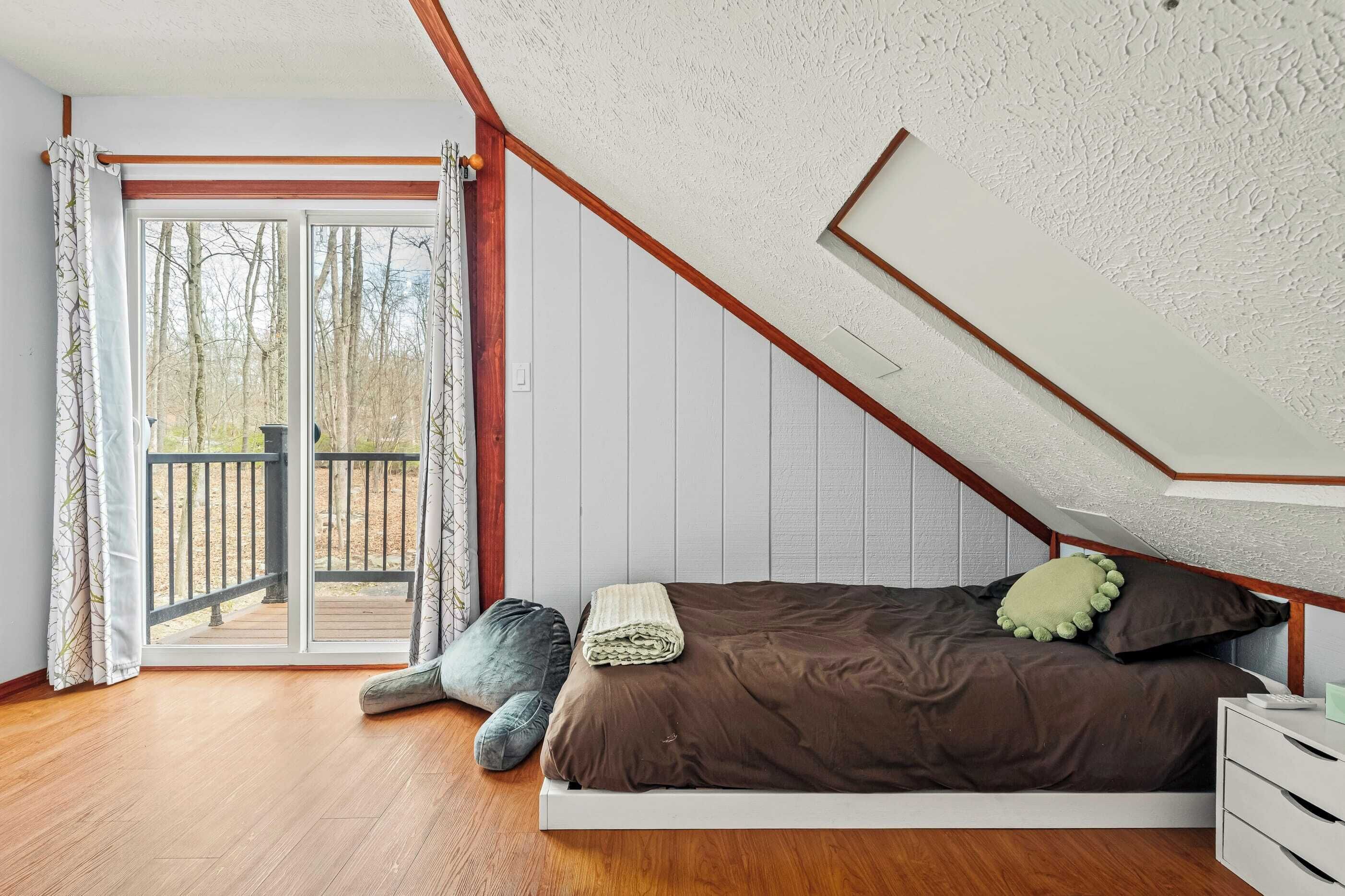 Ein Schlafzimmer mit Holzboden, einem Bett mit brauner Bettwäsche, einem grünen Kopfkissen, einem weißen Nachttisch und Blick durch eine Schiebetür auf eine Terrassenfläche.