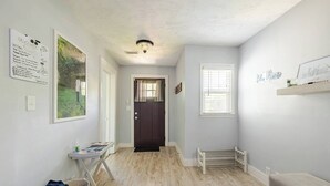 A hallway with a wooden floor, a dark door, a small window with blinds, a white shelf with a sign, and a framed picture on the wall.