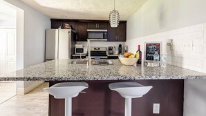 A modern kitchen with dark cabinets, granite countertops, a central island with stools, stainless steel appliances, and a hanging light fixture.