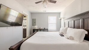 A bedroom with a large bed, a ceiling fan, a TV mounted on the wall, and a window with blinds.