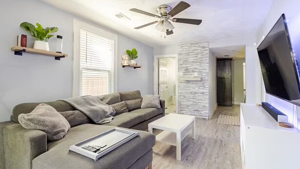 A modern living room with a grey sectional sofa, a white coffee table, a ceiling fan, and a flat-screen TV.
