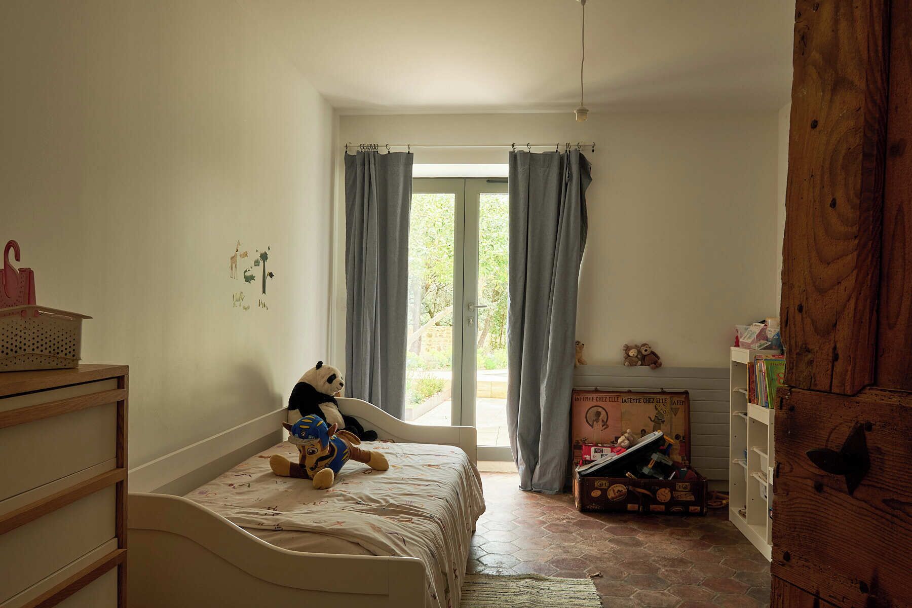 A child's bedroom with a bed, a wooden dresser, a window with curtains, and a bookshelf.
