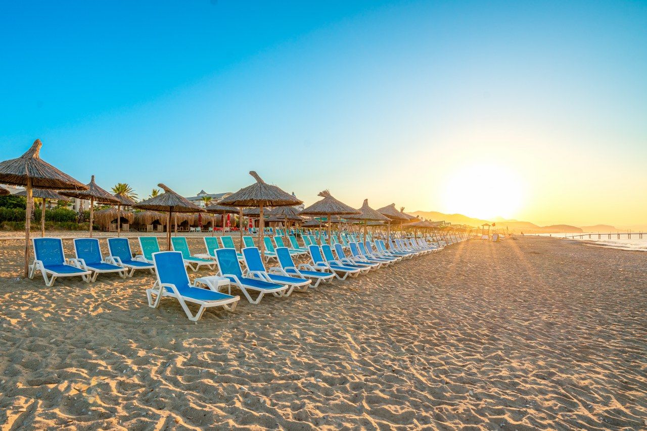 A beach with rows of blue lounge chairs and thatched umbrellas under a clear sky.