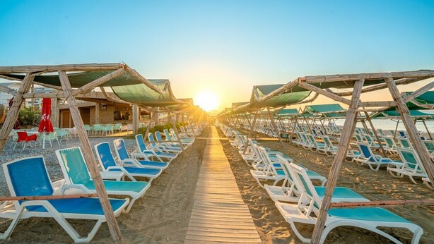 A beach with rows of sun loungers and umbrellas, a wooden walkway, and a building in the background. - Sun Heaven Queen (Alanya)