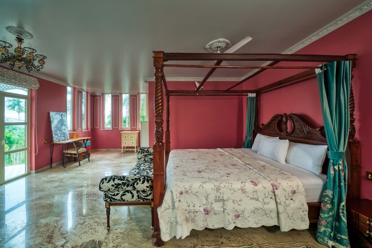 Een ruime slaapkamer met een hemelbed, een houten bureau en een groot raam met uitzicht op palmbomen.