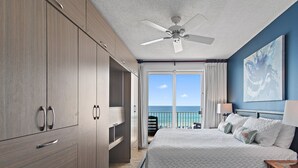 Una habitación de hotel con cama, ventilador de techo, ventana grande con vista al océano y un cuadro en la pared.