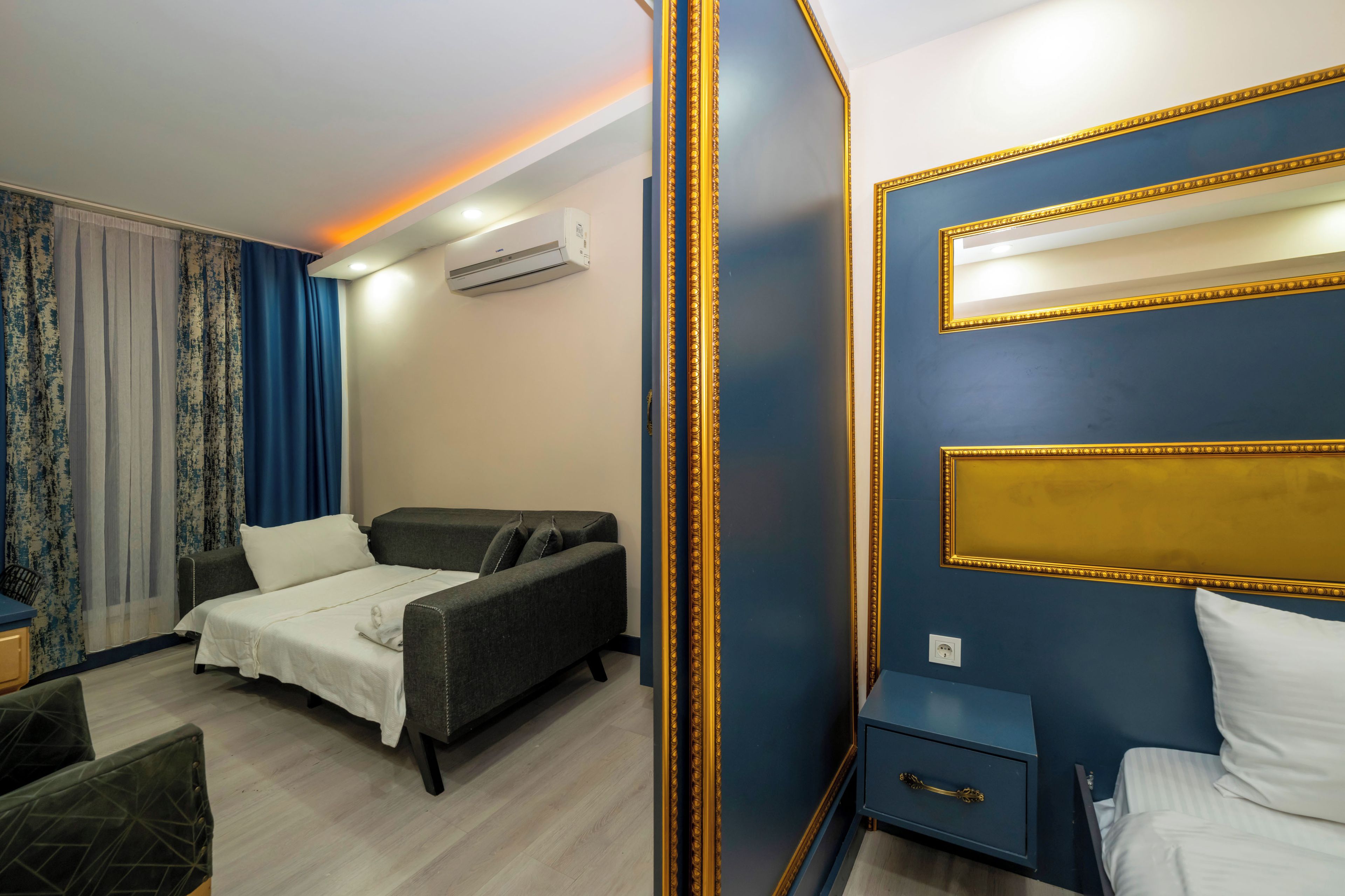 A hotel room with a bed, a sofa, a nightstand, a mirror, and an air conditioning unit.