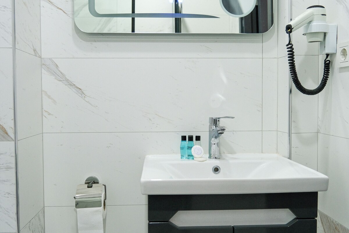A bathroom with a white sink, a mirror, a towel dispenser, and a trash bin.