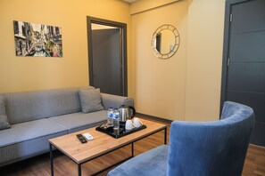 A cozy room with a checkered sofa, a small wooden table with a tray of amenities, a blue armchair, a painting of a canal, and a round mirror on the wall. - La Vitinre (Istanbul)