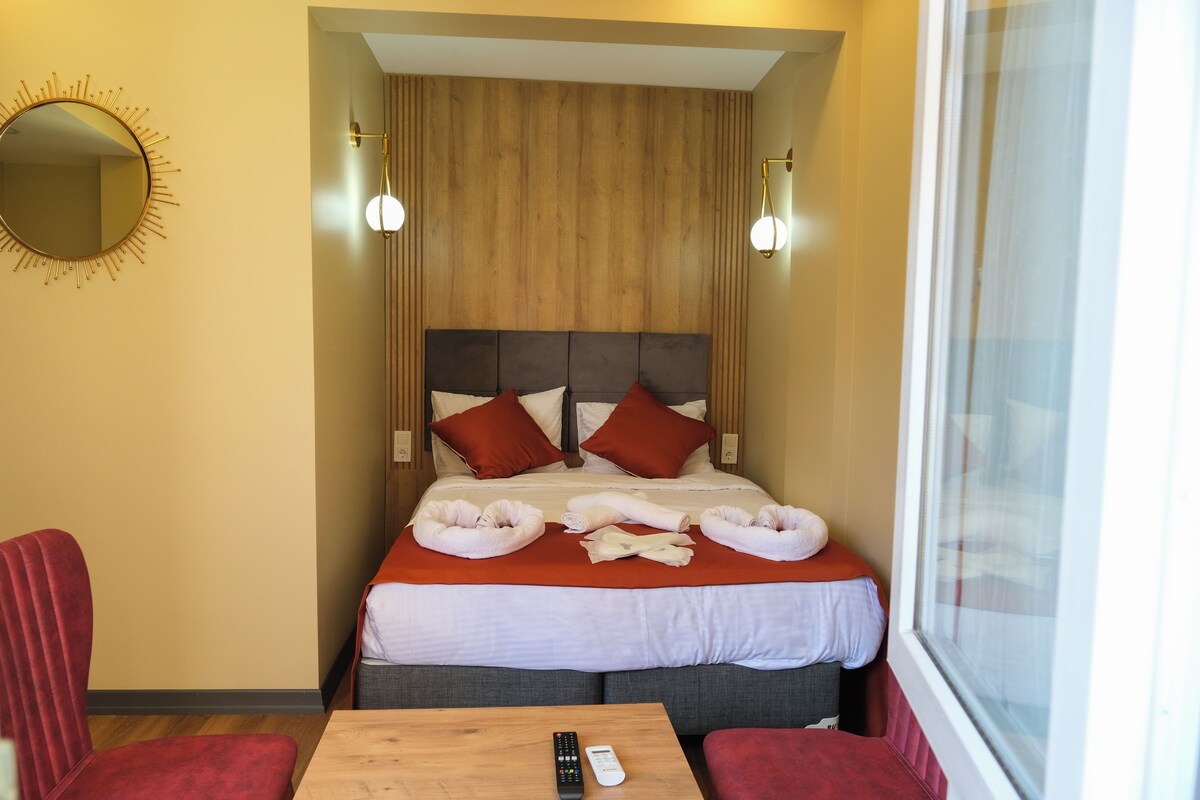 A hotel room with a bed, two red chairs, a wooden table, a remote control, and a window.