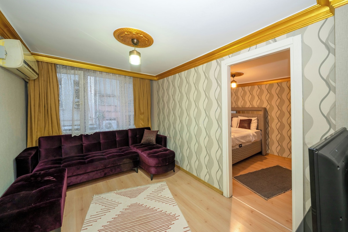 A room with a sectional sofa, a bed, a television, and patterned wallpaper.