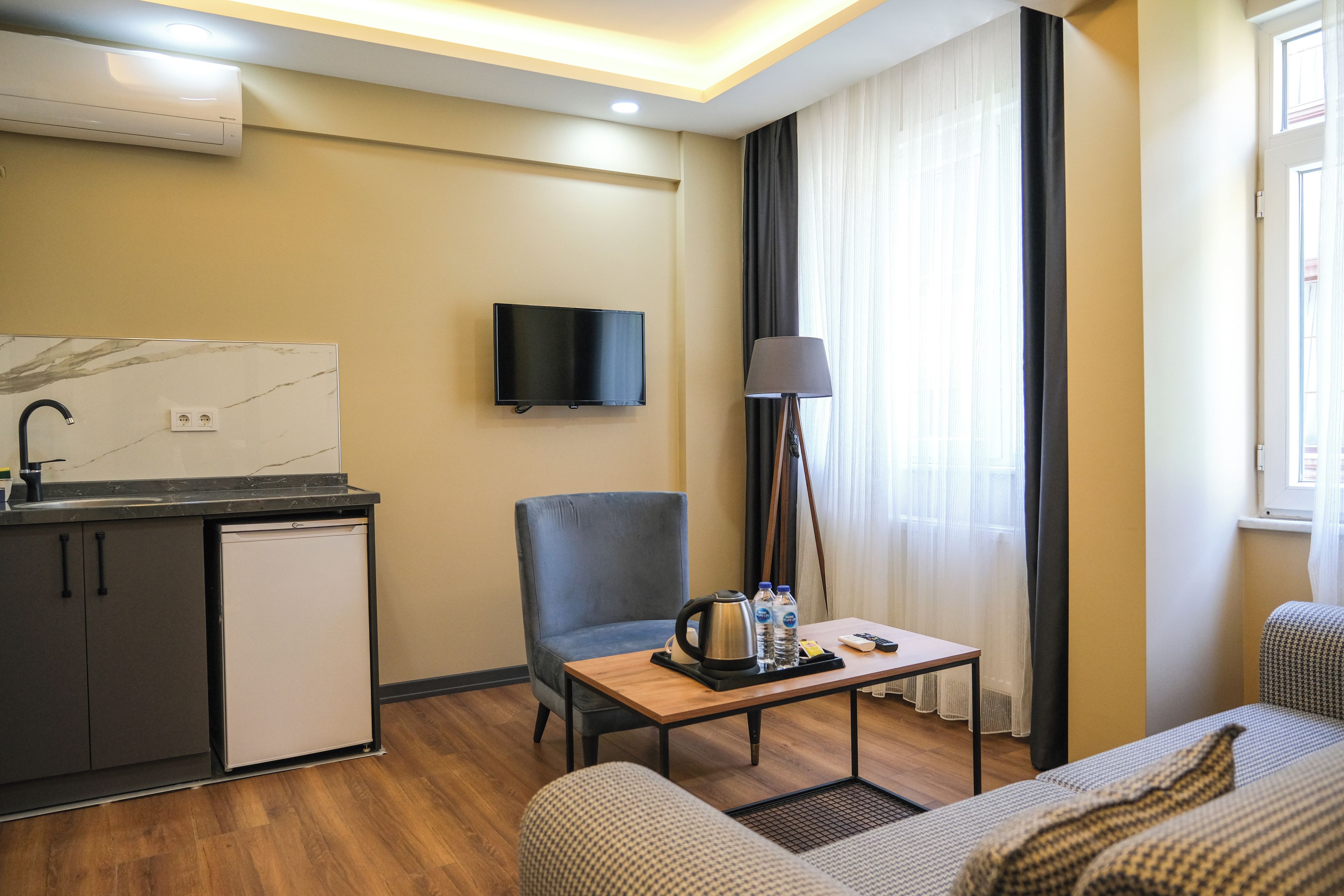 A compact hotel room with a kitchenette, a flat-screen TV, a sofa, a chair, and a table with amenities.