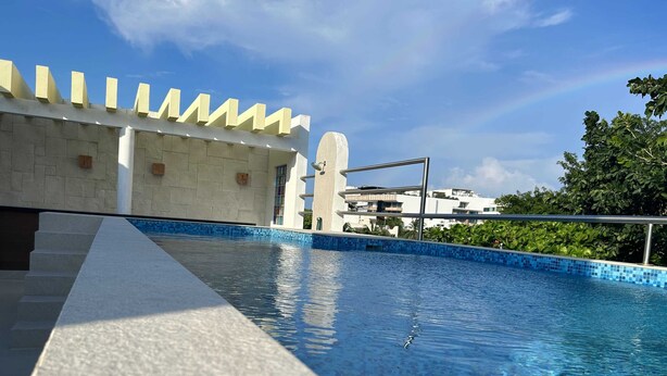A swimming pool with a tiled edge, a stone staircase, and a white building with a unique architectural design in the background. - Tropical Elegance Studio CondoHotel  (Puerto Morelos)