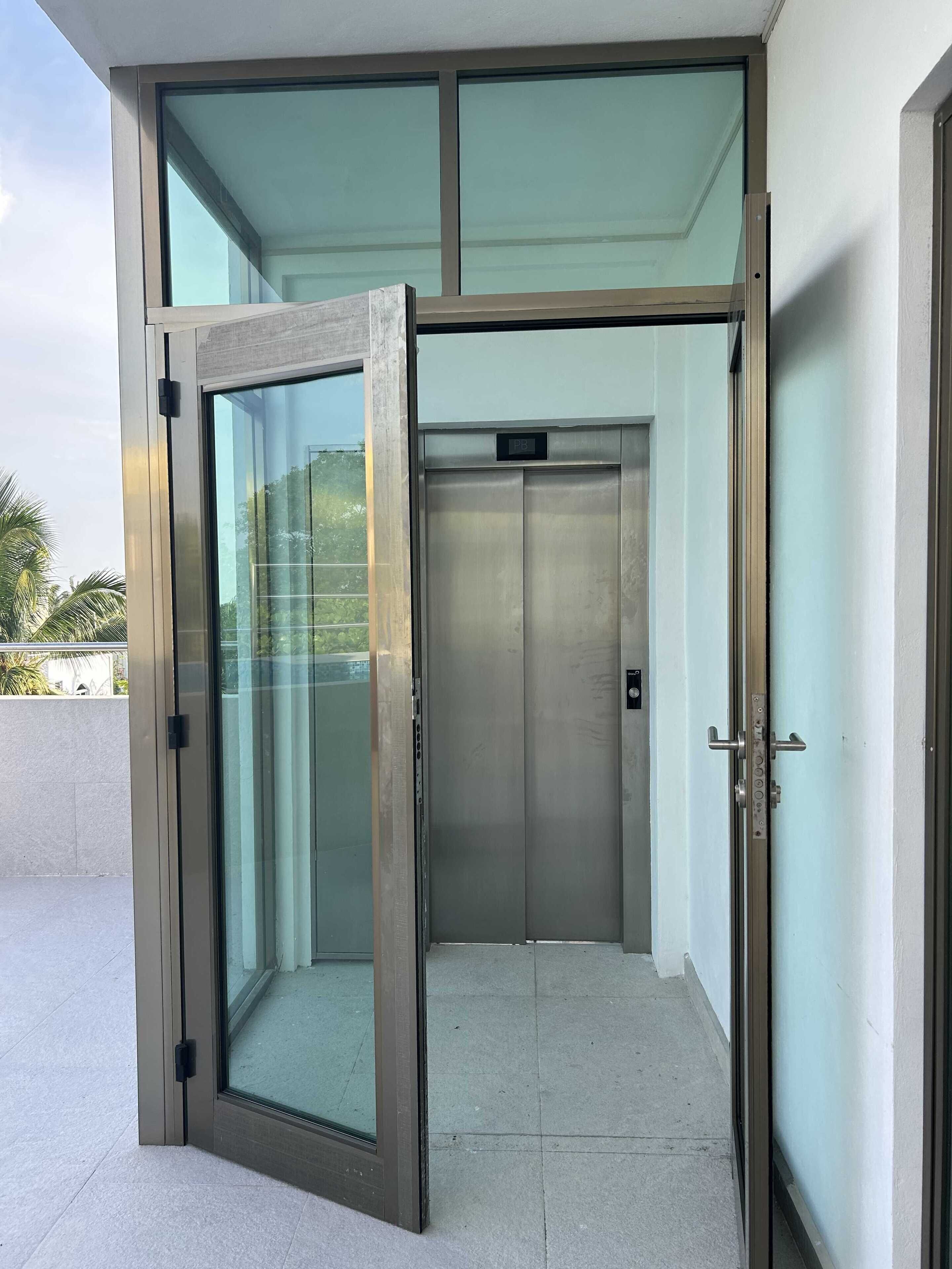 Ascensore con porta in vetro e interni metallici, situato in un edificio moderno con pavimento in cemento e parete di colore chiaro.