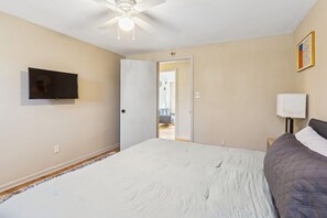 Une chambre à coucher comprenant un lit, un ventilateur de plafond, un téléviseur et une porte donnant sur une autre pièce.