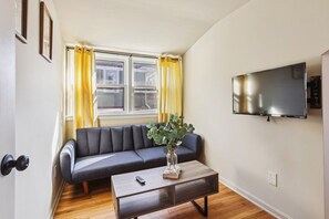 A living room with a sofa, a coffee table with a vase and books, a remote control, a TV on the wall, and a window with curtains.