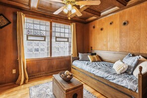 A room with a wooden ceiling, a bed with a blue and white patterned bedspread, a wooden headboard, and a window with decorative glass panes.