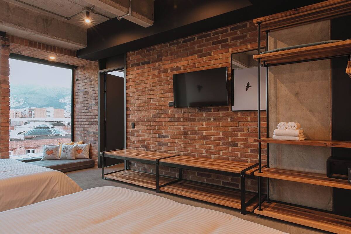 A modern hotel room with a brick wall, a flat-screen TV, a wooden bench, and a bed with a view of the city.