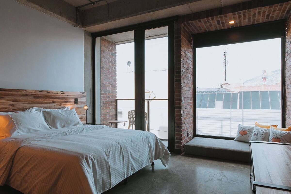 A bedroom with a large bed, a wooden headboard, a balcony with a view of the city, and a small table with chairs.