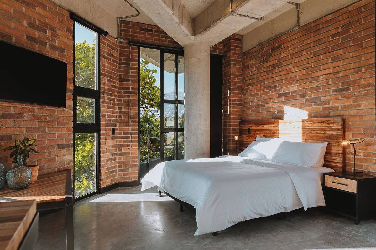 A modern bedroom with a brick wall, large windows, a bed, bedside tables, and a TV mounted on the wall.