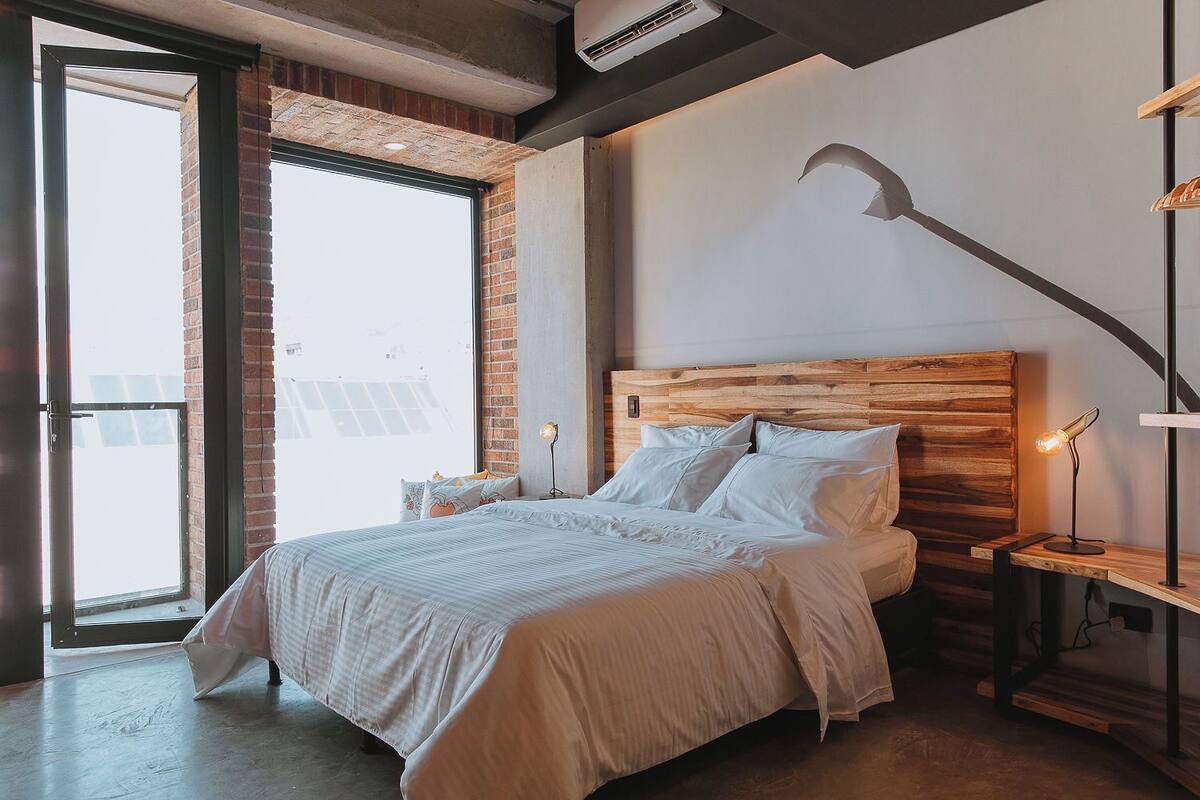A bedroom with a large window, a wooden headboard, a bedside table with a lamp, and a wall-mounted light fixture.