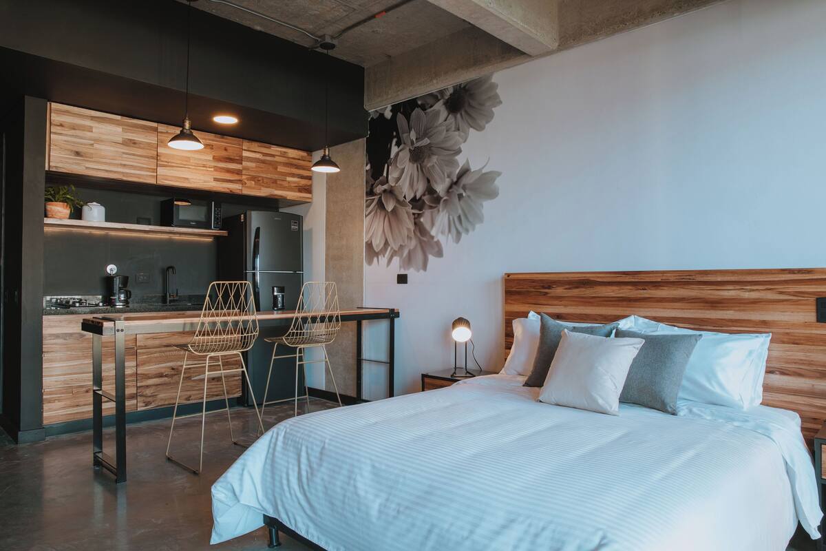 A modern hotel room with a wooden headboard, a large bed, a kitchenette, and a dining area with bar stools.