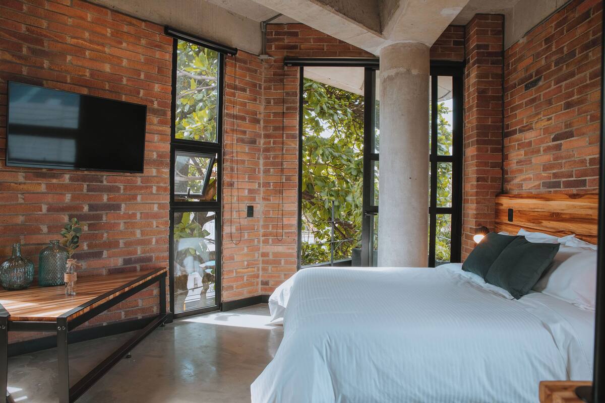A modern bedroom with a brick wall, a large bed, a TV, and a window with a view of trees.