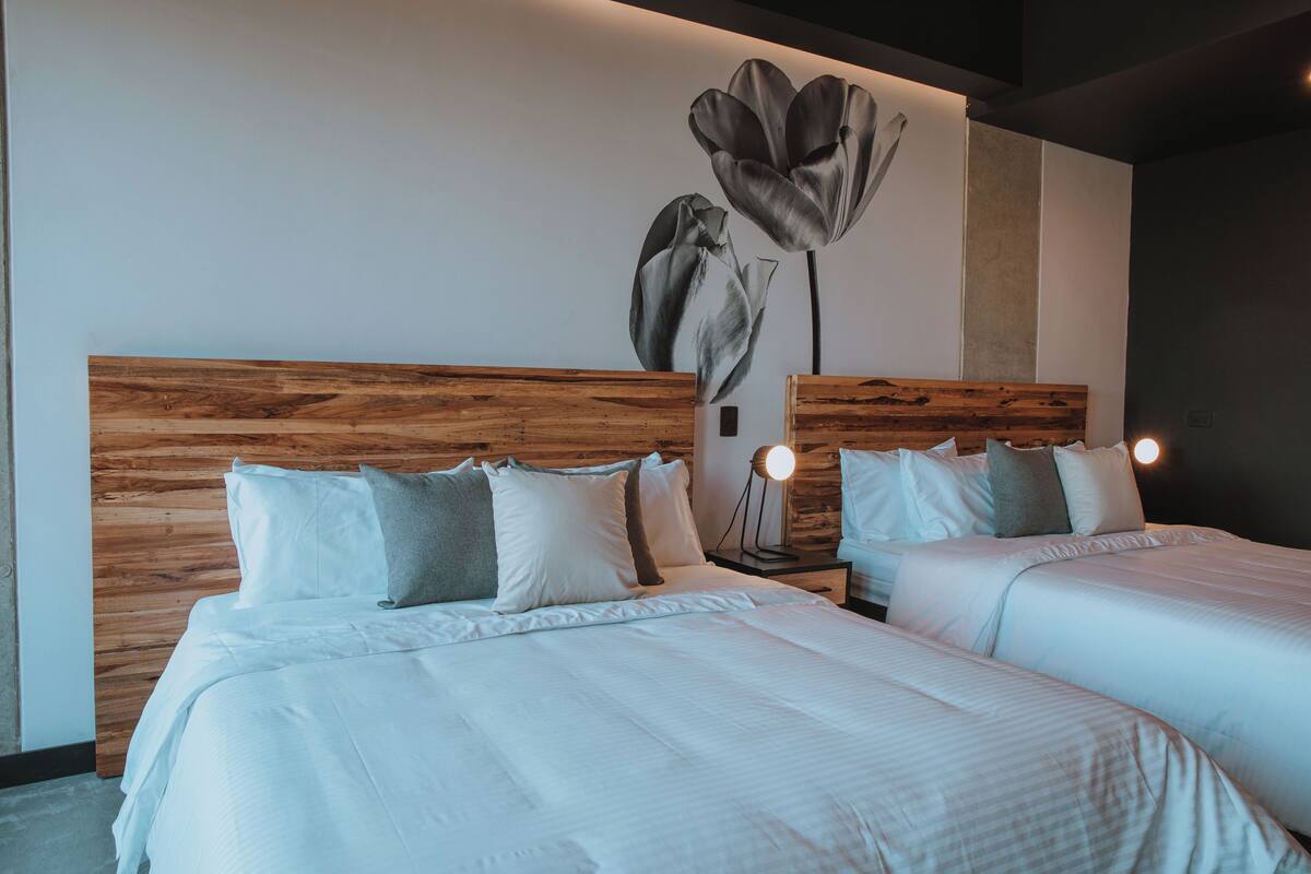 A hotel room with two beds, wooden headboards, white bedding, and a wall-mounted lamp.