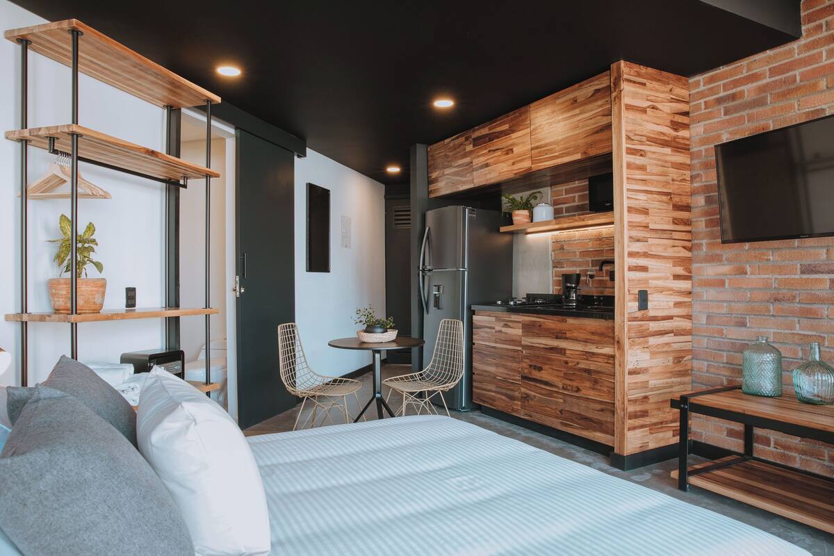 A modern kitchen with wooden cabinetry and a built-in TV. There is a dining area with a table and chairs, and a bed with pillows.