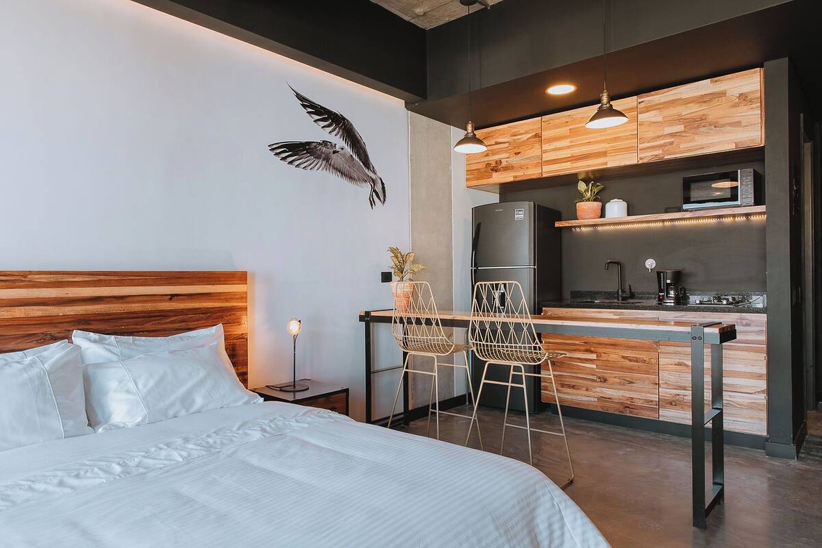 A modern bedroom with a wooden headboard, a bed with white linens, a nightstand with a lamp, and a kitchenette with a microwave, stove, and bar stools.