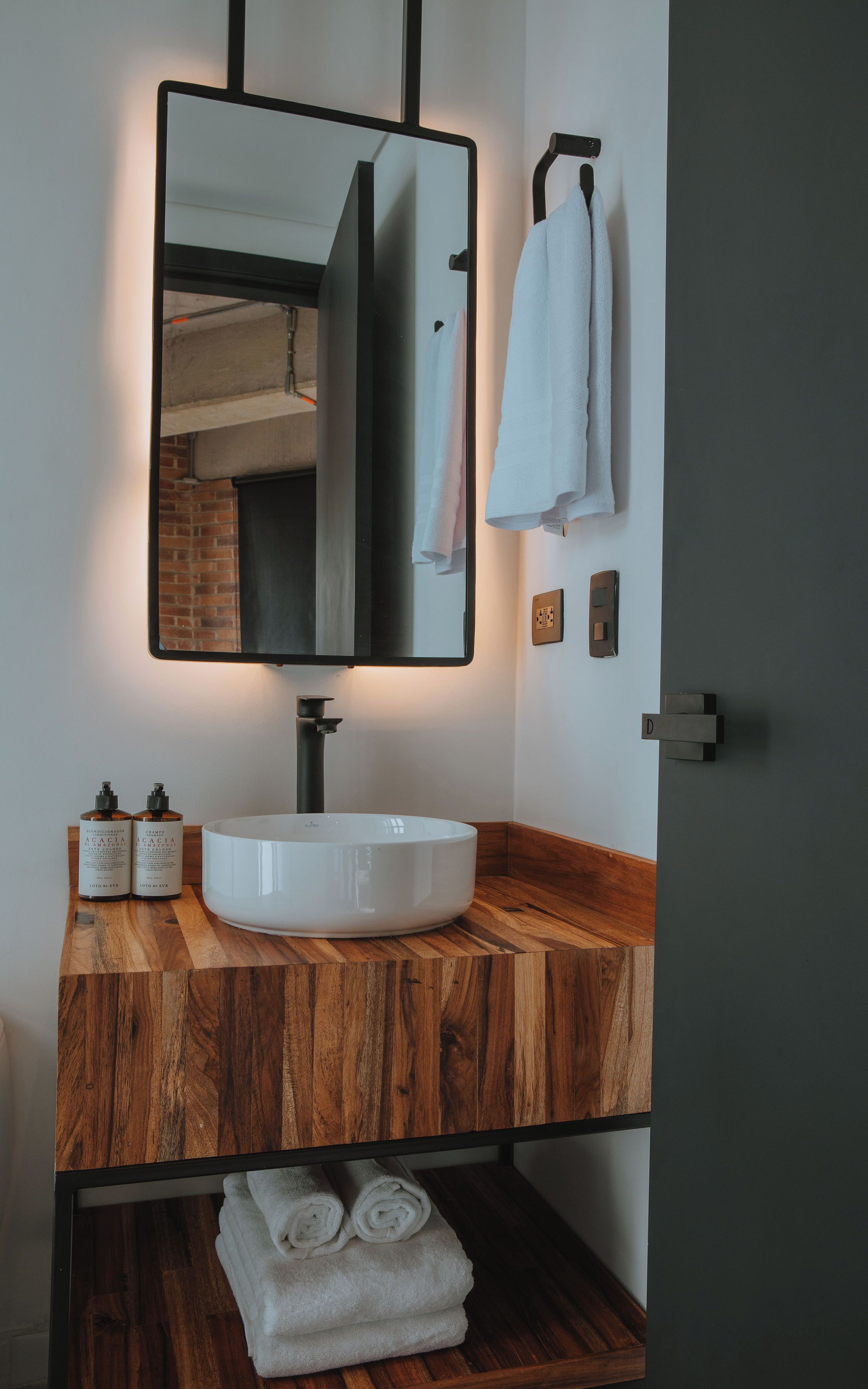 A bathroom with a wooden vanity, a white sink, and a mirror. There are two bottles of shampoo on the vanity, and a towel is hanging on the wall.